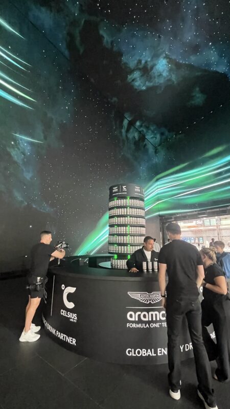 Aston Martin Aramco and Celsius event stand with a tower of energy drink cans and attendees under a starry display